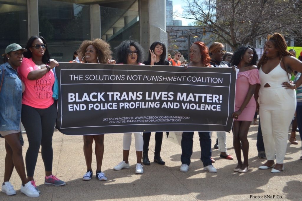 Outdoors during the daytime, a row of Black women hold and gather round a black banner with white text, which reads: 