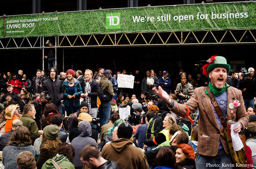 A crowd sits in the middle of a downtown street during Occupy Toronto. A man in a plaid blazer and green hat rabble-rouses. A TD Bank banner overhead reads: 