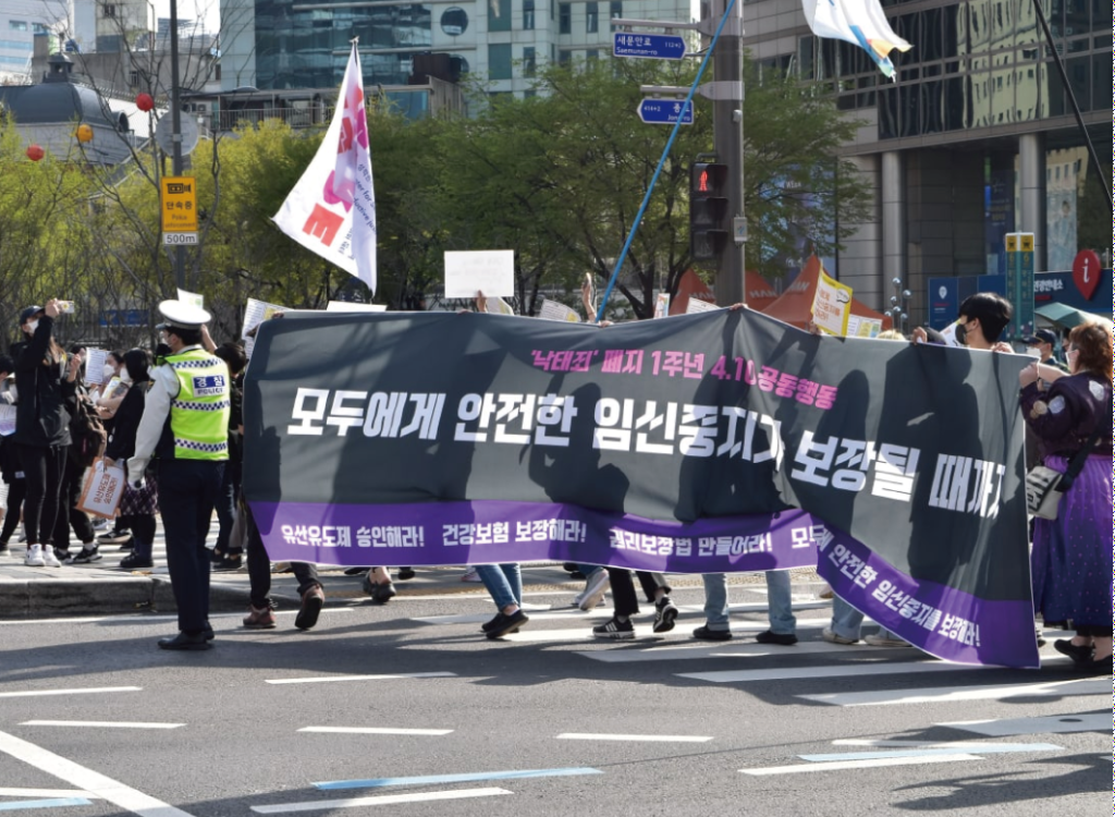 A picture of a rally on April 10, 2022. The participants are holding a placard displaying the sentence (in Korean), "Until safe abortion is assured for everyone." At the bottom of the placard, other demands: approve Mifegymiso, expand national health insurance to cover every case of abortion, legislate the Framework Act on the Guarantee of Sexual and Reproductive Rights.