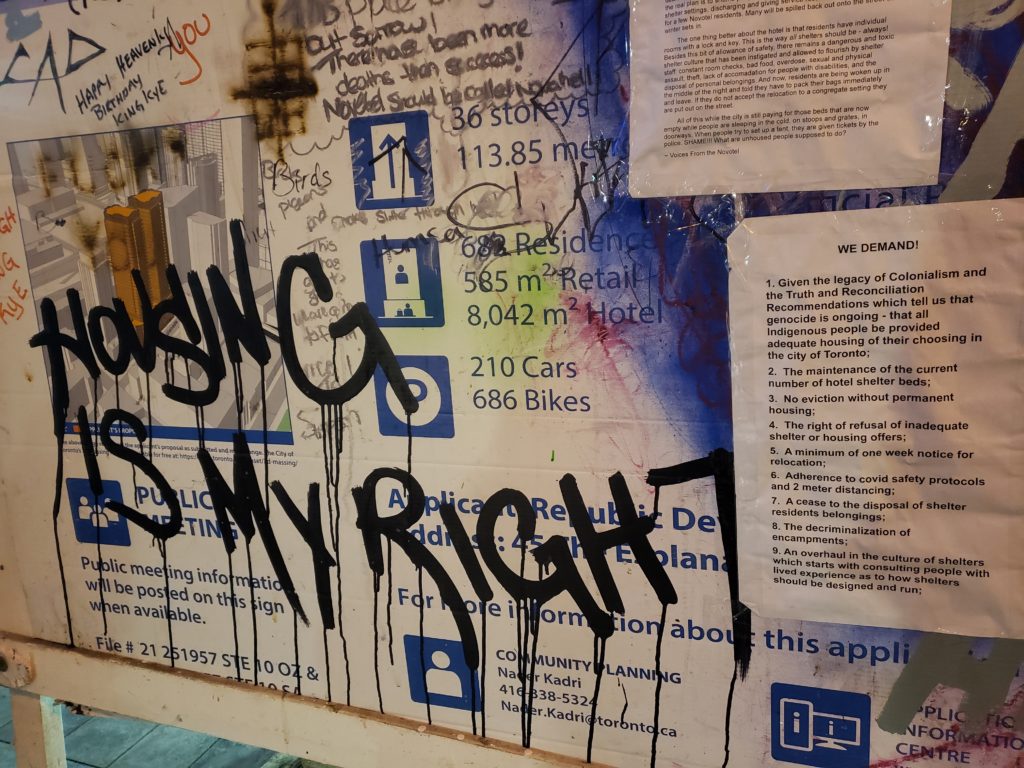 A city notice-of-development billboard is covered with graffiti that reads "Housing Is My Right" and other markings, along with a posted sheet of paper that lists housing demands.