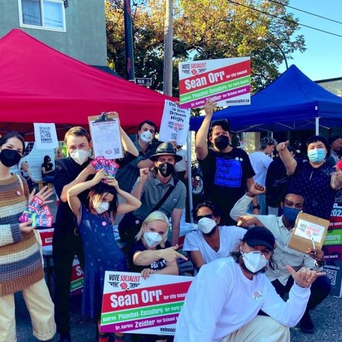 Supporters of the VOTE Socialist campaign, nearly all wearing respirator-type masks, pose at an outdoor campaign event. The sky is blue and makeshift campaign tent-pavilions are set up in the background. The supporters hold up campaign signs and clipboards.