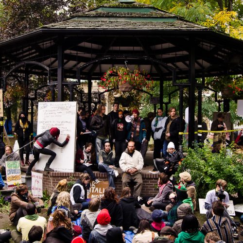 A few dozen people gather under and beside a gazebo in St. James Park during Occupy Toronto. There are some placards; one person writes on a white board; someone else holds a megaphone.