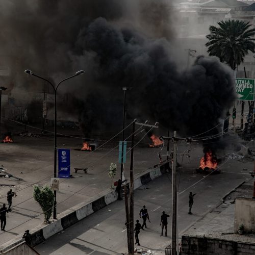Black smoke plumes above a broad street in Lagos, Nigeria, where police with guns drawn are scattered. A palm tree is visible amidst buildings with corrugated metal roofs.