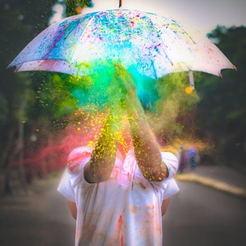 A photo of a person, or maybe two people back-to-back, standing in a road and holding a white umbrella as splashes of paint, in queer and trans pride flag colours, explode all over it.