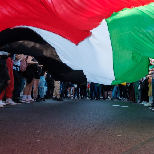 A crowd, a collective, stands around and holds up a giant Palestine flag. Only their legs, feet, shoes can be seen.