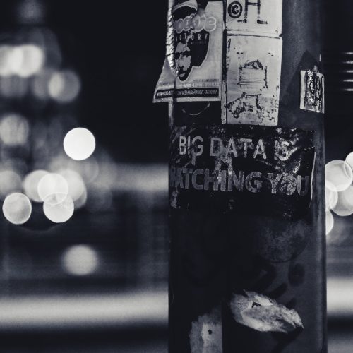 A black and white photo of a city street with twinkling nights. Pasted on a pole, a sign reads "Big Data is Watching You."