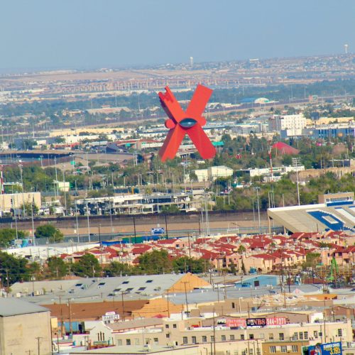 A large red X sculpture in Juarez, Mexico as seen from downtown El Paso, Texas, USA. Both cities are dense urban environments with low structures; many of the houses have red rooftops. The sky is a hazy blue-grey.