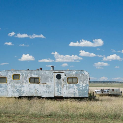 A rusted trailer and car flipped upside down, set against a plateau and blue sky speckled with clouds.
