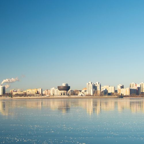 A photo of the Volga River in Kazan, Tatarstan. The sky is blue, the water shining; residential high-rises and industrial buildings dot the shoreline.