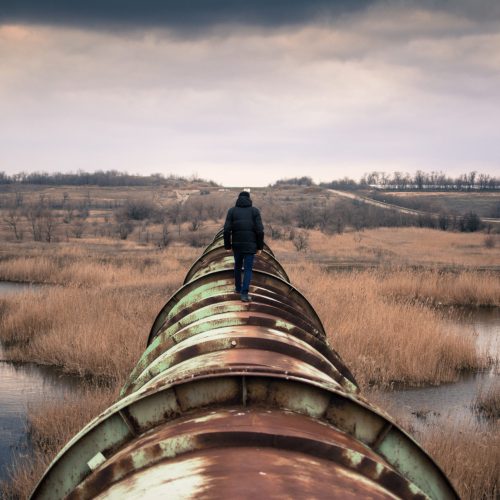 A solitary figure walks along a big rusted pipe cutting across the land, including a stream or river. The sky is dramatic with thick clouds, and the land dry but lovely.